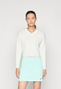 Λευκή κοντή μακρυμάνικη μπλούζα με V-λαιμόκοψη, συνδυασμένη με μια ανοιχτή μέντα πράσινη φούστα που διαθέτει το μαύρο λογότυπο της Nike στο στρίφωμα. Λαμπερή υφή, απλός σχεδιασμός.