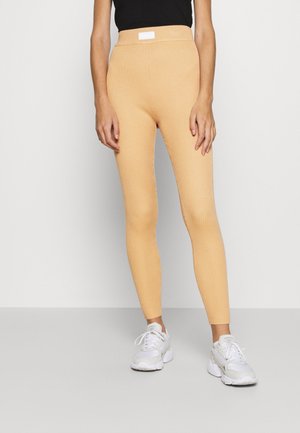 Femme portant des leggings côtelés beiges, un top noir sans manches et des baskets blanches, debout devant un fond clair uni.