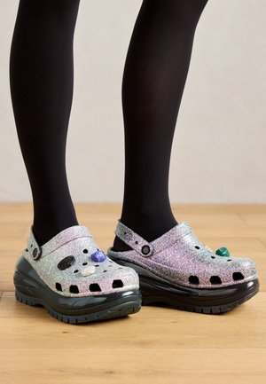 Clogs argentés pailletés avec des semelles noires, ornés de gemmes décoratives, comprenant des trous de ventilation ouverts et une sangle sur le dessus pour un ajustement sécurisé.