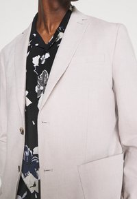 Blazer de lino color beige claro con cierre de un solo botón, solapas de muesca y un bolsillo en el pecho. Se lleva sobre una camisa de estampado floral negro.