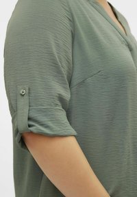 Chemise légère vert olive avec une texture froissée, manches courtes et un détail à roll-up avec boutons. Elle présente une poche discrète à l'avant.