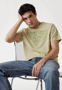 Bežinis medvilnės marškinėliai su žalia grafinė spauda, derinami su mėlynomis džinsinėmis kelnėmis. Modelis sėdi skaidriame krėsle, remiasi ranka į galvą.