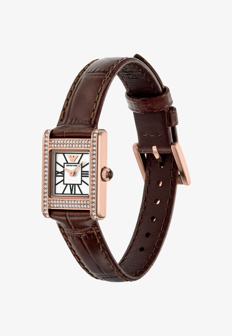 Obdelhové hodinky ve růžovém zlatě s bílým ciferníkem, černými římskými číslicemi, korunkou zdobenou krystaly a hnědým koženým řemínkem s přezkou.