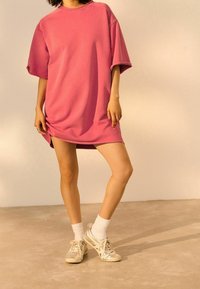 Roze oversized sweatshirt jurk met korte mouwen. Zachte stof, relaxte pasvorm en een zoom boven de knie. Draagt met zilveren sneakers en witte sokken.