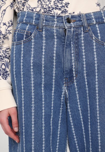 Blue denim jeans with vertical white stripes, a straight cut, five pockets, and a button-fly closure. The fabric has a textured finish.