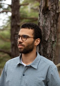 Sinine ja valge ruuduline nööpidega särk, kraega; ümmargused kilpkonnakujulised prillid; näokarvad; seisab puu kõrval.