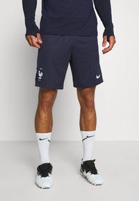 Mężczyzna w granatowych sportowych szortach z białym symbolem koguta i logo Nike, w białych skarpetach Nike oraz czarno-białych korkach stoi na białym podłodze.
