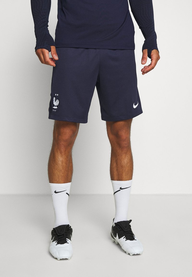 Mężczyzna w granatowych sportowych szortach z białym symbolem koguta i logo Nike, w białych skarpetach Nike oraz czarno-białych korkach stoi na białym podłodze.