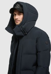 Tumedat puffer-jakk featuring kõrge krae, suur kapuuts ja elastsed detailid. Jakk sisaldab logo kleebist varrukal ja tõmblukku.