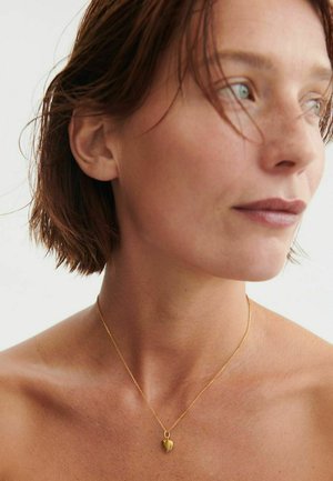 Donna con capelli corti castani che indossa una delicata collana a catena d'oro con un piccolo ciondolo a forma di cuore, che guarda di lato.