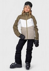 Jaunas žmogus vilki trigubos spalvos slidinėjimo striukę, juodas slidinėjimo kelnes, juodas pirštines, pilkus slidinėjimo batus, pilką beanie skrybėlę ir juodus slidinėjimo akinius ant galvos.