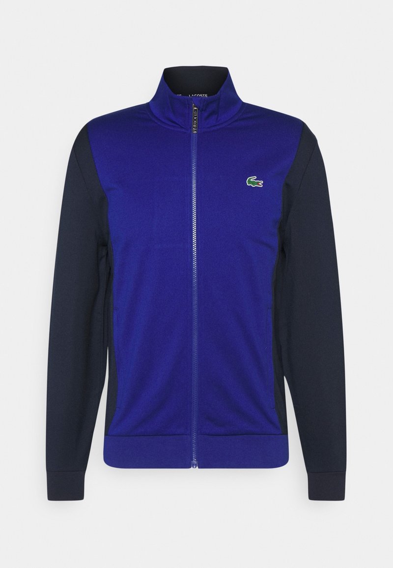Blaue Zip-Jacke mit marineblauen Ärmeln, aus glattem Material. Mit kleinem grünen Krokodil-Logo auf der Brust. Zwei Taschen an den Seiten.