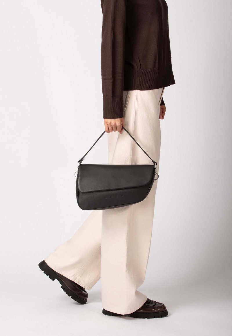 Černá kožená kabelka s zakřiveným designem, vybavená krátkým popruhem a kovovými doplňky. Modelka má na sobě světle zbarvené široké kalhoty.