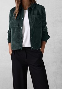 Dark green corduroy jacket with button closure, two chest pockets, and a relaxed fit, worn over a white t-shirt and black wide-leg pants.
