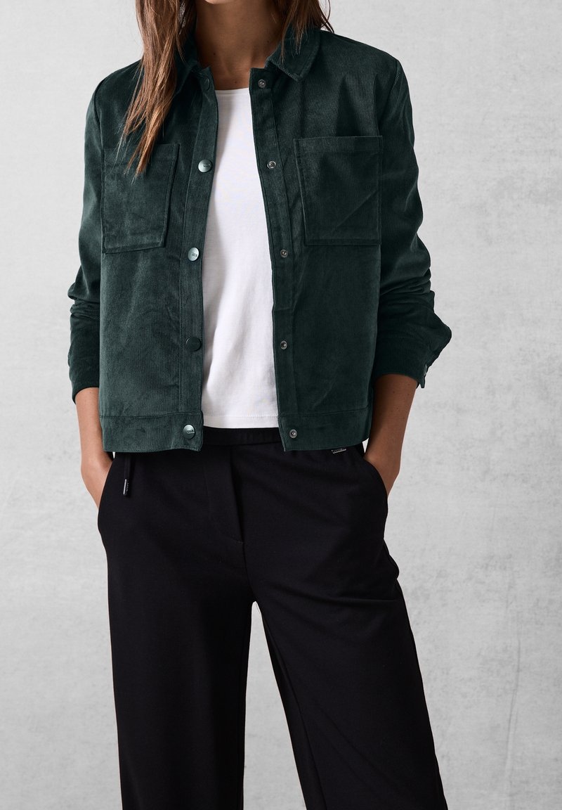 Dark green corduroy jacket with button closure, two chest pockets, and a relaxed fit, worn over a white t-shirt and black wide-leg pants.