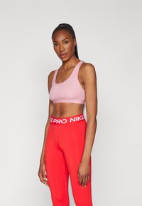 Roza sportska podložna majica s širokim trakama i bijelim Nike logom, u paru s crvenim tajicama koje imaju bijeli Nike Pro pojas.