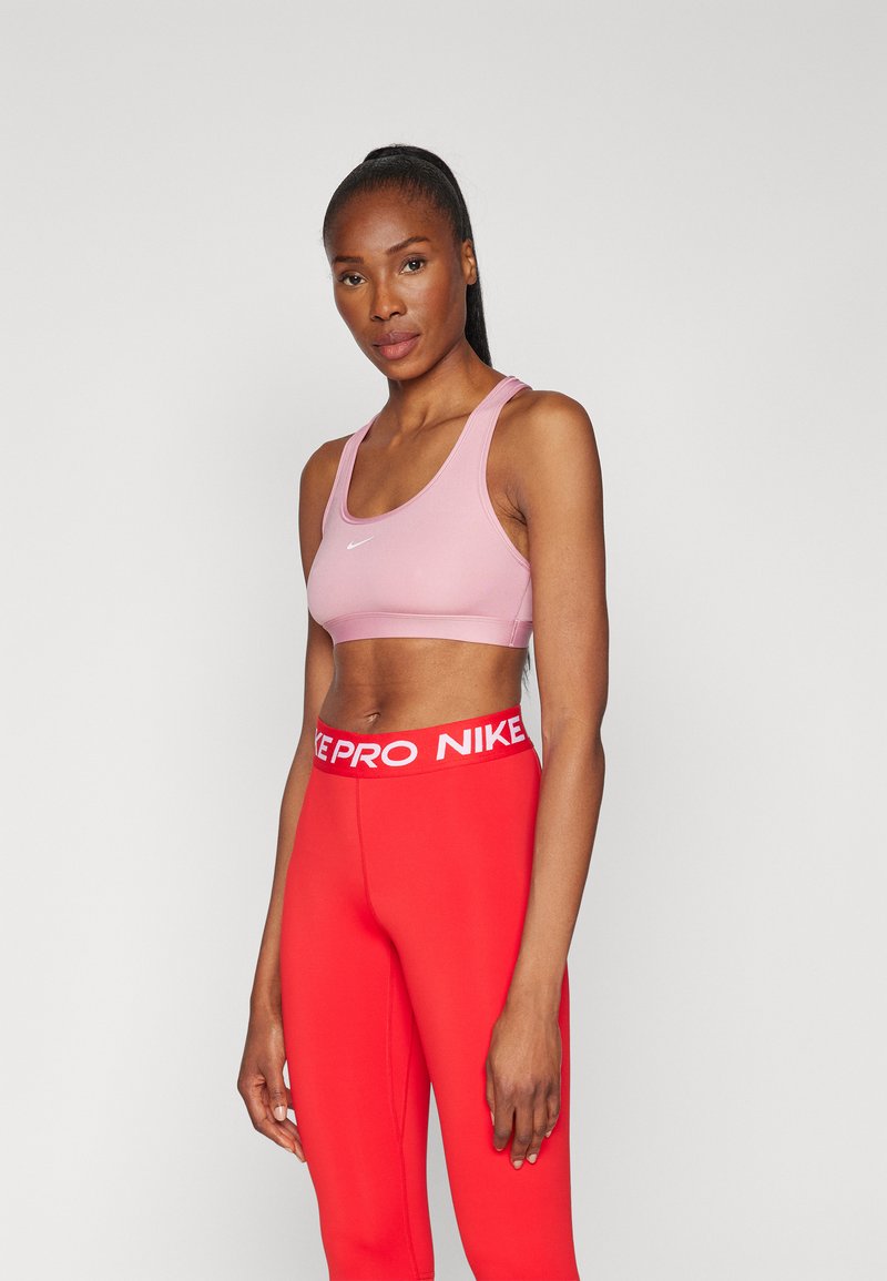 Roza sportska podložna majica s širokim trakama i bijelim Nike logom, u paru s crvenim tajicama koje imaju bijeli Nike Pro pojas.