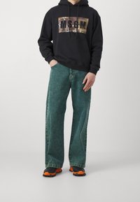 Czarna bluza z kapturem z grafiką w kamuflażu i napisem "MSGM"; zestawiona z luźnymi zielonymi jeansami oraz czarnymi sneakersami z pomarańczowymi akcentami.