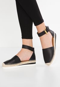 Espadrille de cuero negro con tira al tobillo y hebilla dorada, con suela de yute trenzado y suela de goma blanca.