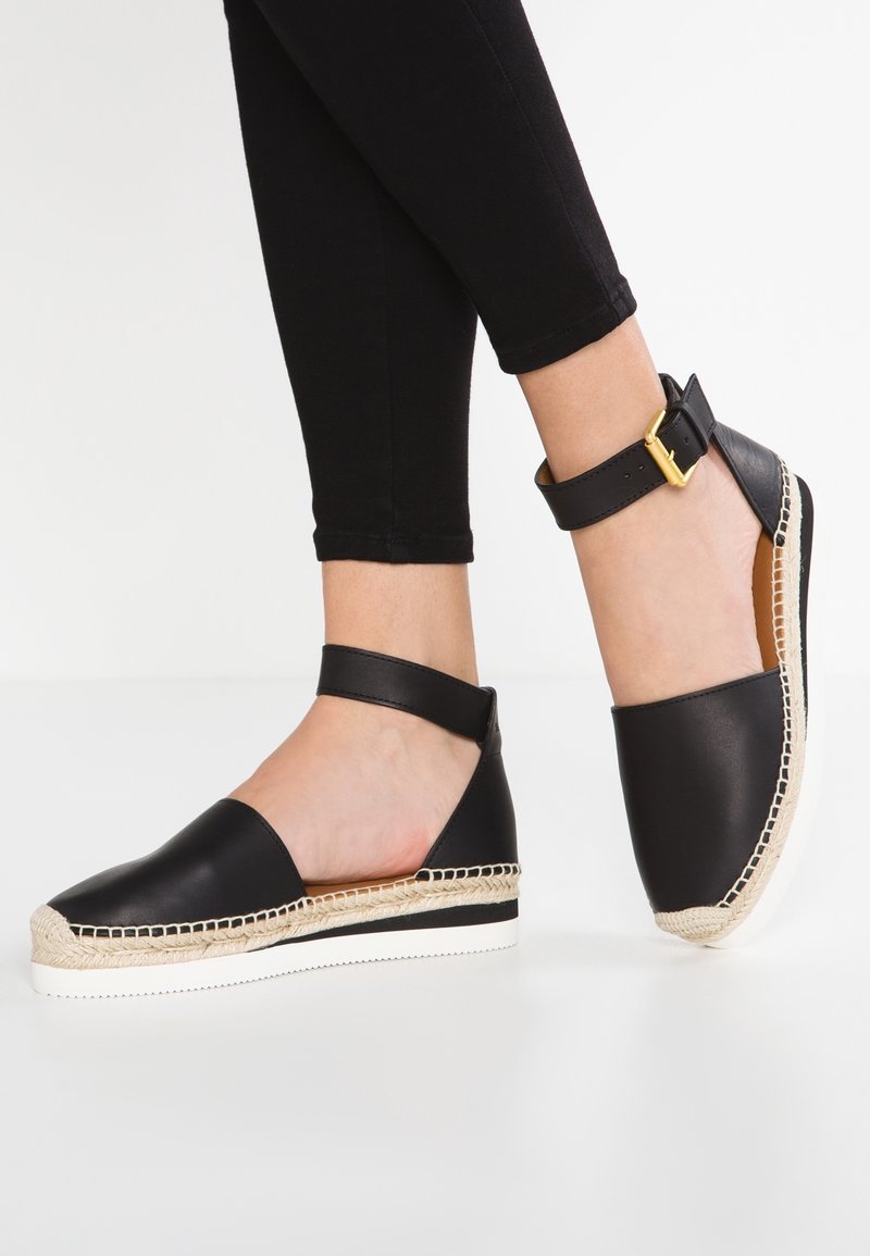 Espadrille de cuero negro con tira al tobillo y hebilla dorada, con suela de yute trenzado y suela de goma blanca.
