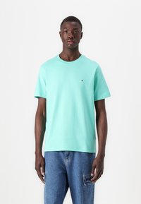 Lyse mintgrønn t-skjorte i bomullsmateriale, med rund hals og korte ermer, prydet med en liten logo på brystet, sammen med blå denimjeans.