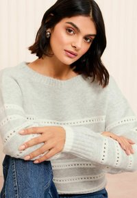 Pullover tricotat de culoare gri deschis cu dungi orizontale albe, având detalii cu ochiuri și manșete riblate. Este purtat cu blugi, cu brațele încrucișate.