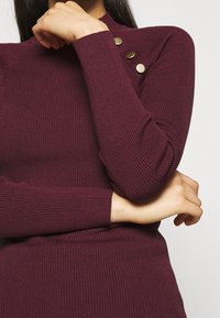 Maglia a maniche lunghe bordeaux a coste con un collo alto e tre bottoni dorati sull spalla, dal taglio aderente.