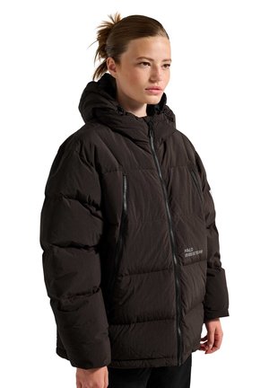 Junge Person mit braunen Haaren zu einem Dutt gebunden, trägt eine schwarze Daunenjacke mit Kapuze und Reißverschlusstaschen, schaut leicht zur Seite.