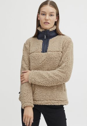 Pull en molleton beige avec un col zippé bleu marine. Tissu extérieur texturé, coupe décontractée et poche poitrine insérée avec un logo de marque discret.