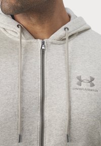 Ingrandimento di un uomo che indossa un hoodie zip-up grigio chiaro di Under Armour con cordini, che mostra il collo, la cerniera e il logo sul petto.