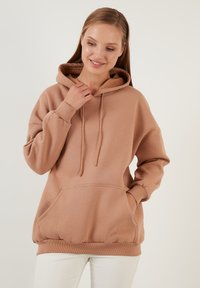 Przytulny, beżowy sweatshirt w oversize'owym kroju, wykonany z miękkiego materiału. Posiada przednią kieszeń, regulowany kaptur na sznurki oraz ściągacze przy rękawach i u dołu.