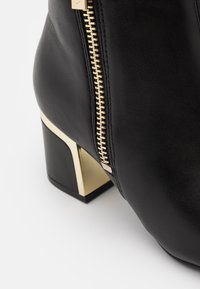 Bota de cuero negro hasta el tobillo con detalle de cremallera metálica dorada, tacón cuadrado bloque con un acento dorado, textura suave y forma estructurada.