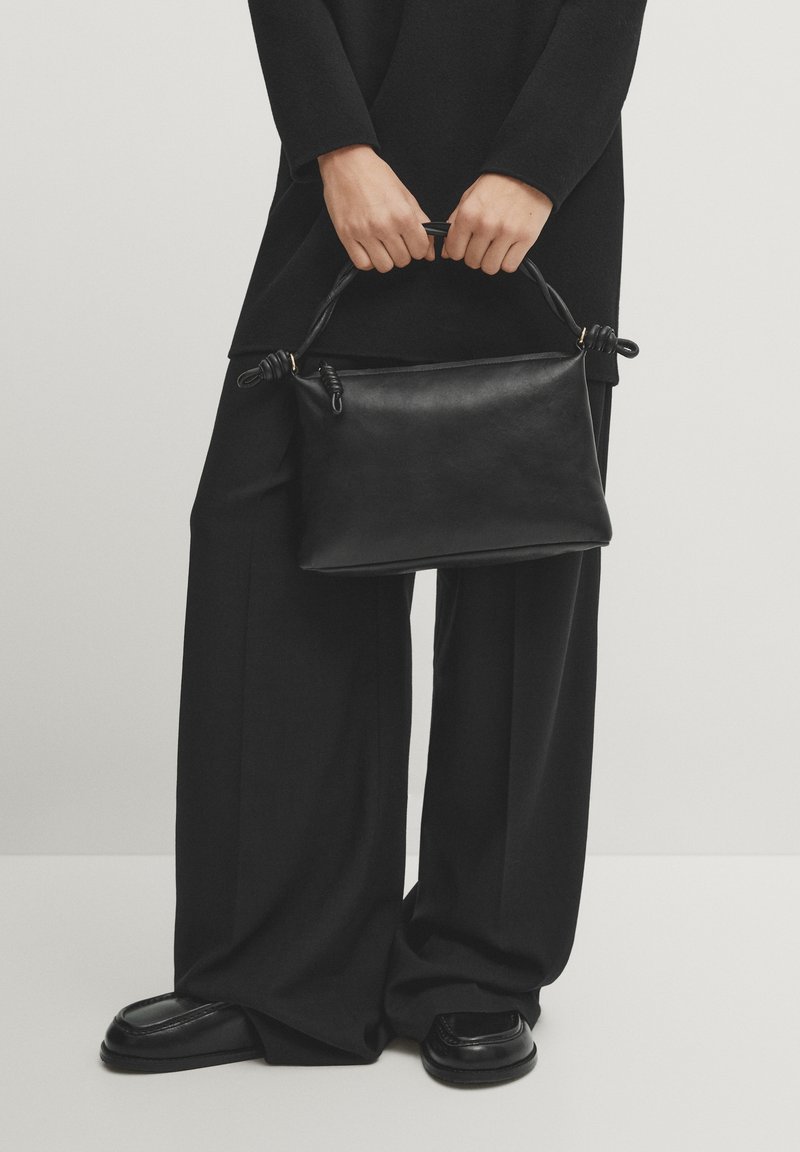 Personne portant un pantalon large noir et des chaussures tient un sac à main en cuir noir avec une poignée torsadée devant un fond uni.