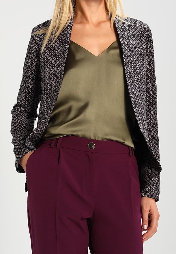 Femme portant un blazer noir à motifs, un caraco en satin vert olive et un pantalon taille haute couleur prune avec les mains dans les poches.