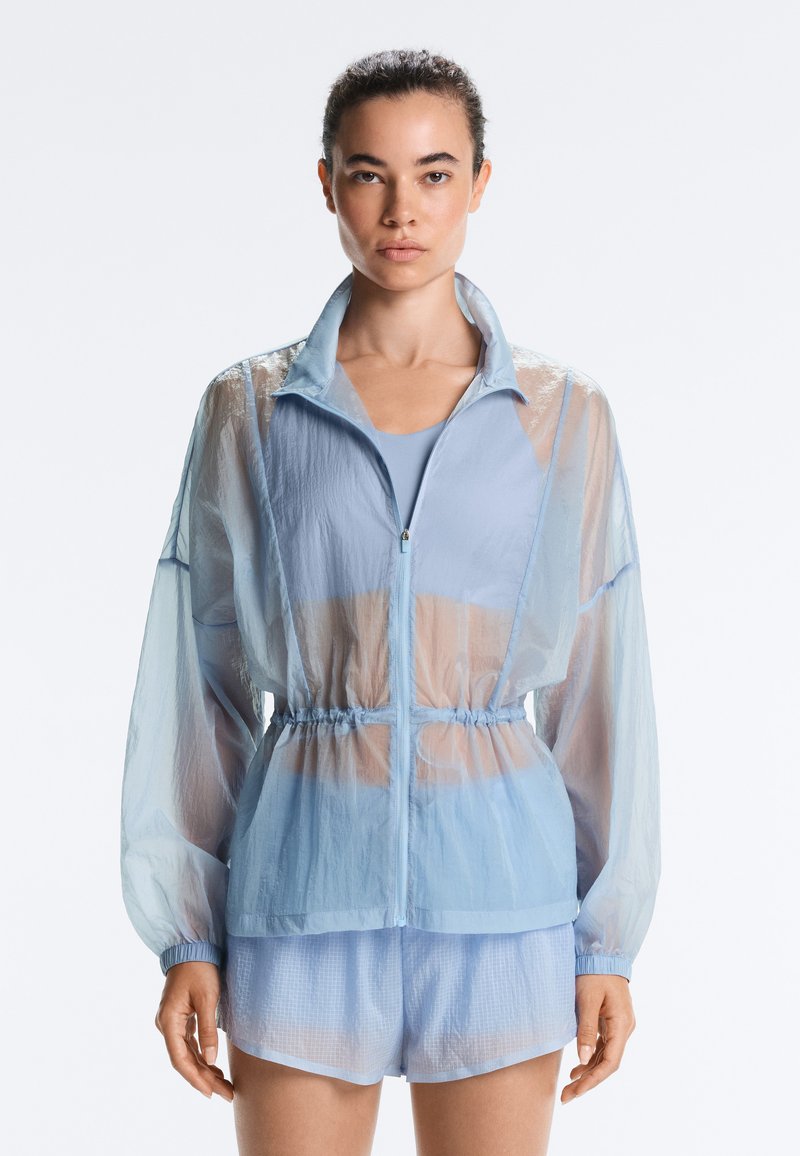 Femme portant une veste zippée légère bleu clair transparente avec taille élastique et short assorti, debout devant un fond clair uni.