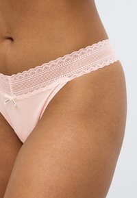 Roze tanga met een delicaat kanten taillebandje met een gegolfde rand en een kleine strik aan de voorkant. Zachte en gladde textuur.