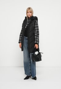 Svart polstret vest med hette, lagd over en svart topp og rutete skjorte. Brede denimjeans og svart håndveske fullfører antrekket.