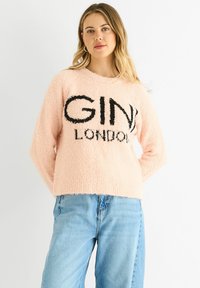 Pink fluffy sweater featuring black "GINI LONDON" text, round neckline, long sleeves, and a cropped fit. Worn with light blue denim jeans.