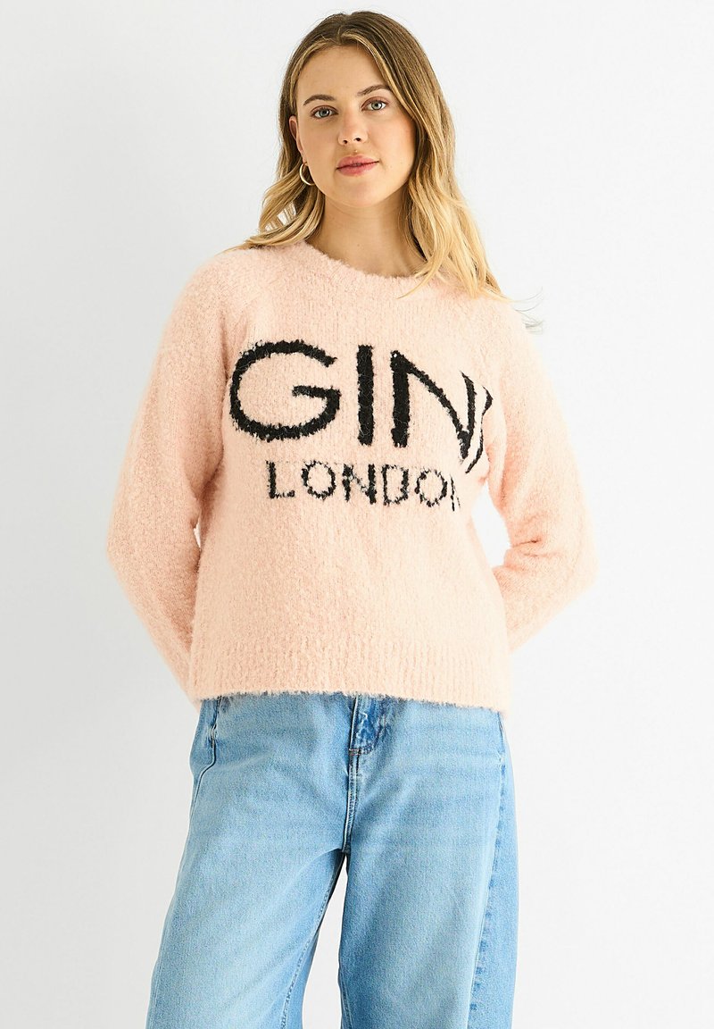 Pink fluffy sweater featuring black "GINI LONDON" text, round neckline, long sleeves, and a cropped fit. Worn with light blue denim jeans.