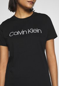 Černé bavlněné tričko s krátkými rukávy, se výrazným bílým logem "Calvin Klein" na hrudi. Hladká textura, klasický střih.