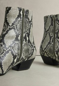 Tekstúrájú, kígyóbőr mintás bokacsizmák műbőrből, amelyeket cipzáras záródás és jellegzetes fekete tömbsarkak jellemzik.