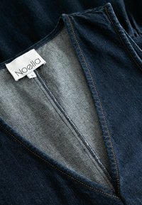 Denim krekls ar V veida dekoltē, tumši zilā krāsā, pelēku oderi, oranžām šuvēm un ar birku, uz kuras rakstīts "Noella" un izmērs "S".