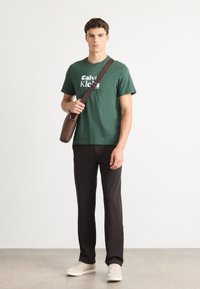 Young man standing in black pants, green Calvin Klein t-shirt, beige sneakers, holding a brown crossbody bag strap over shoulder.