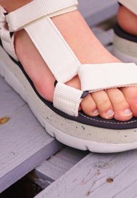 Vita tygremmar sandaler med en tjock, fläckig sula. Remmarna säkrar över foten. Tårna är delvis synliga, med en slät hudstruktur.