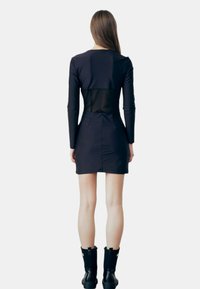 Zwarte aansluitende jurk, lange mouwen, doorzichtige achterkant. De stof lijkt glad met een elegante, op maat gemaakte silhouet. Gecombineerd met zwarte enkellaarsjes.