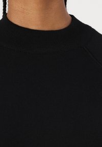 Melnas adītas džemperis ar ribotu apļa kaklu. Auduma tekstūra izskatās maiga un gluda, ar piegrieztiem piedurkņu piegriezumiem un pieguļošu siluetu.