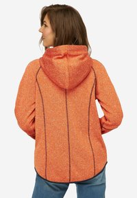 Felpa arancione in tessuto texturizzato con cappuccio, caratterizzata da cuciture scure a contrasto e orlo curvo, indossata sopra jeans blu.