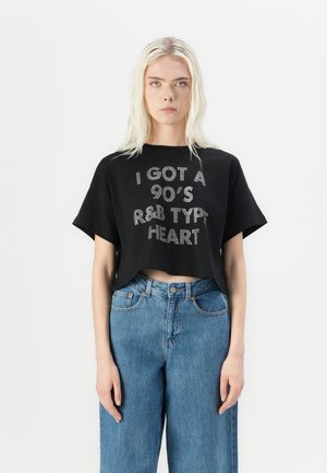 Black cropped t-shirt with silver rhinestone text "I GOT A 90’S R&B TYPE HEART," paired with high-waisted blue jeans. Plain background.