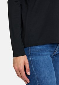 Långärmad svart skjorta i mjukt tyg, tillsammans med blå jeans. Skjortan har en enkel design med rak skärning och fåll.