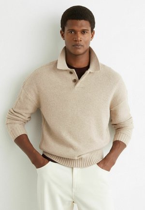 Homme portant un pull en tricot beige avec un col relevé et un pantalon blanc, debout les mains dans les poches contre un mur blanc.
